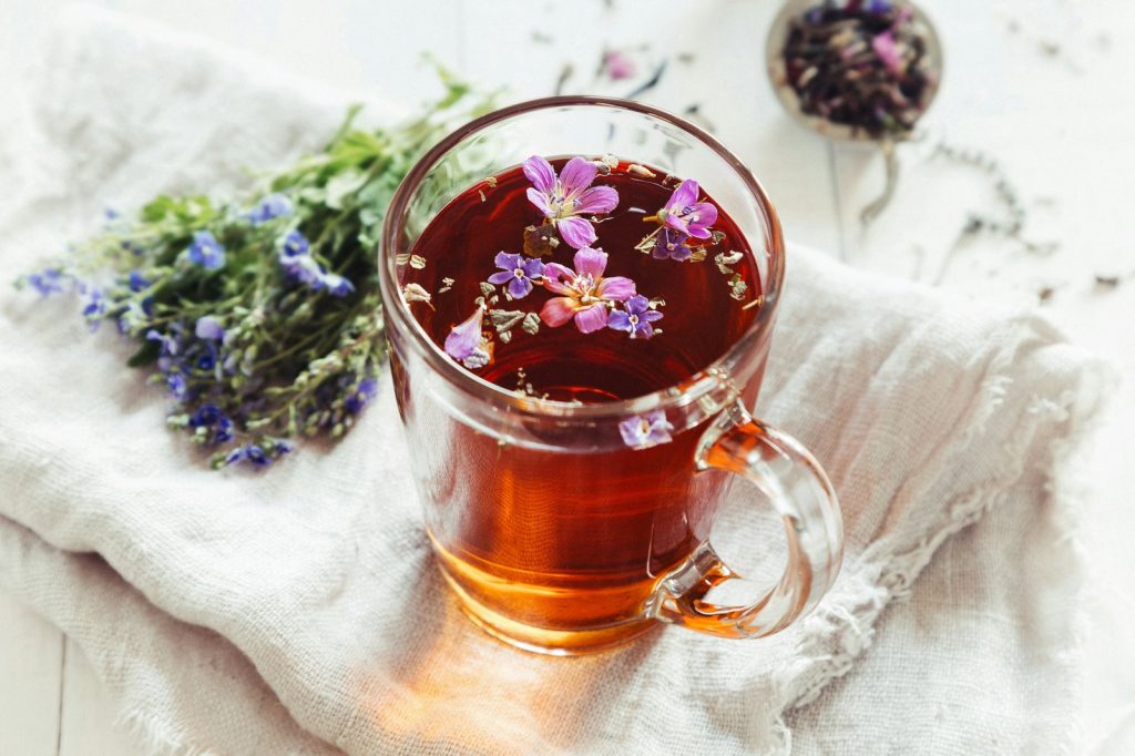 Glas mit Kräutertee und kleinen Blüten, daneben frische Kräuterzweige mit blauen Blüten auf einem hellen Leinentuch
